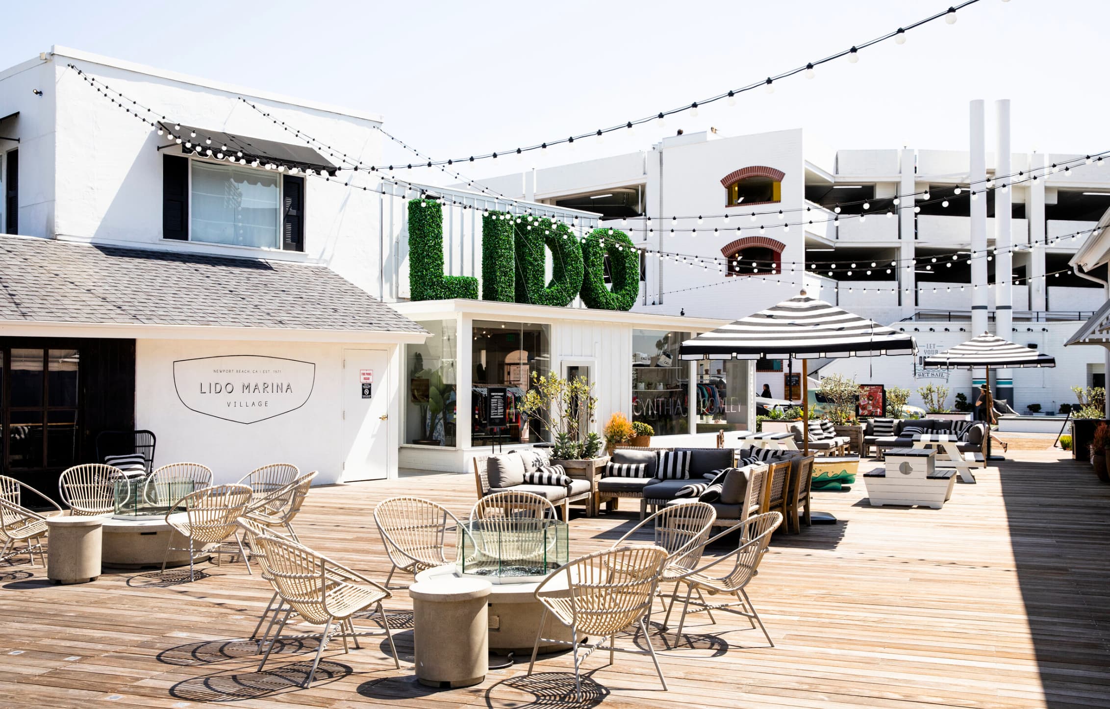 Lido Marina Village boardwalk in Newport Beach — green topiary LIDO sign, string lights, and waterfront lounge seating.